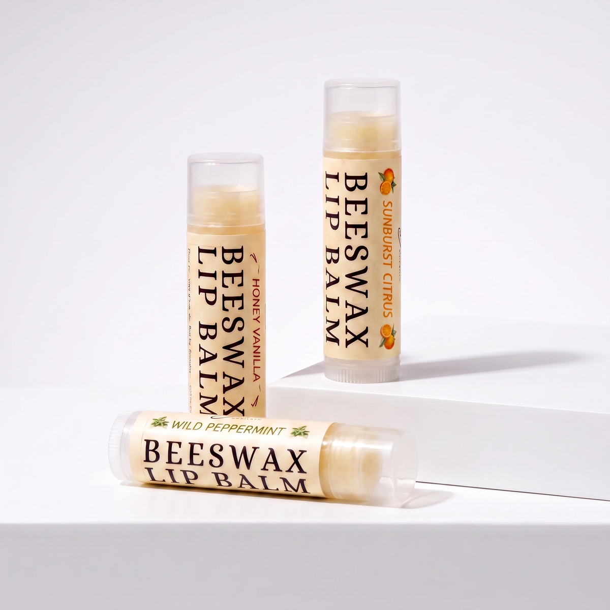 Three tubes of beeswax lip balm arranged on a white surface against a white background. One tube is lying down in front reading 'Wild Peppermint,' and two tubes stand up behind reading 'Beeswax Lip Balm' with 'Sunburst Citrus' on the right and 'Bee Sweet Vanilla' on the left.